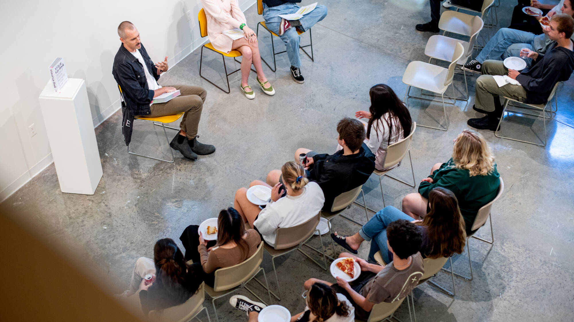 A group of people sit in a circle in a spacious room, engaging in conversation while holding plates of food.